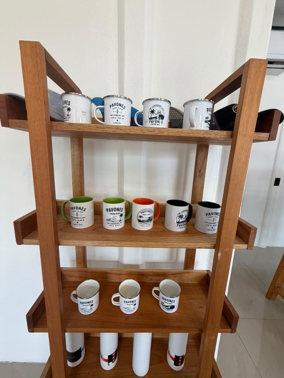 Wooden shelf of Pavones Surf Club enamel mugs, ceramic mugs and water bottles