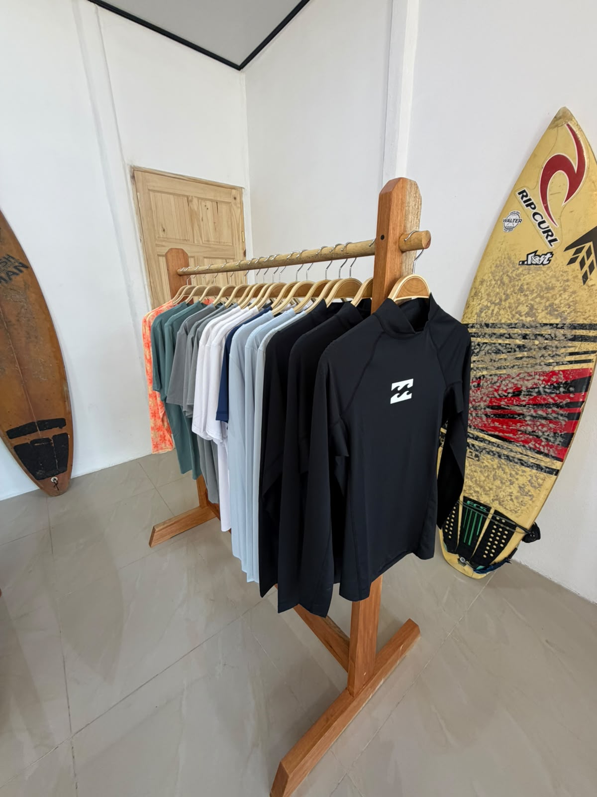 Rack of Billabong rash guards and surf tees inside Pavones Surf Club, with a Rip Curl surfboard leaning on the white wall