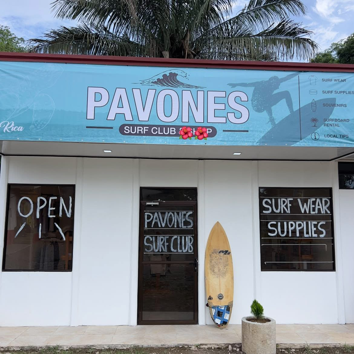 The Pavones Surf Club storefront: white building with a blue Pavones awning, PAVONES SURF CLUB lettering on the glass door and a surfboard leaning outside
